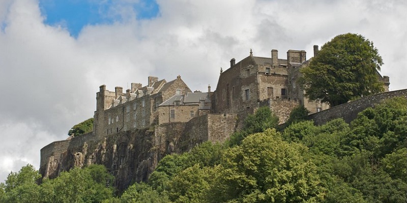 Photo of Stirling Castle against a blue and slightly cloudy sky, and surrounded by greenery.