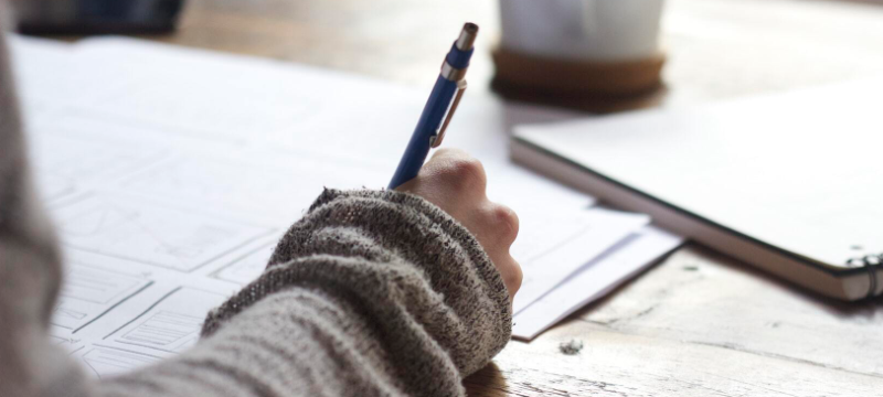 Image of someone writing with a pen. Paper and a notebook are pictured on the table.