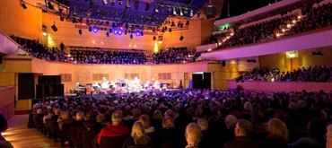 A photo of a theatre hall with a stage at the far end and people seated in the foreground 