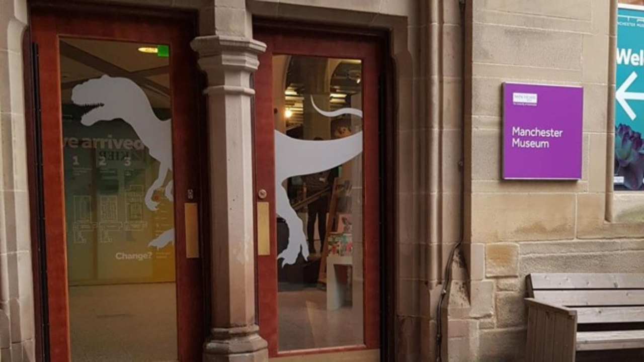 The accessible entrance of the Manchester Museum, a Victorian Gothic-style building with arched windows and ornate stonework. The entrance is marked by large wooden doors.