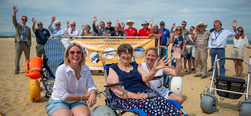 a picture of someone in a beach wheelchair on Ryde beach with two volunteers kneeling beside them. In the background there are other volunteers and council officials weaving from behind a banner.