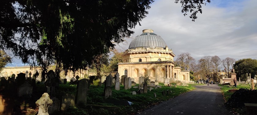 A historic cemetery with rows of headstones and ornate mausoleums. A stone path leads through the grounds, which is lined with trees and greenery.