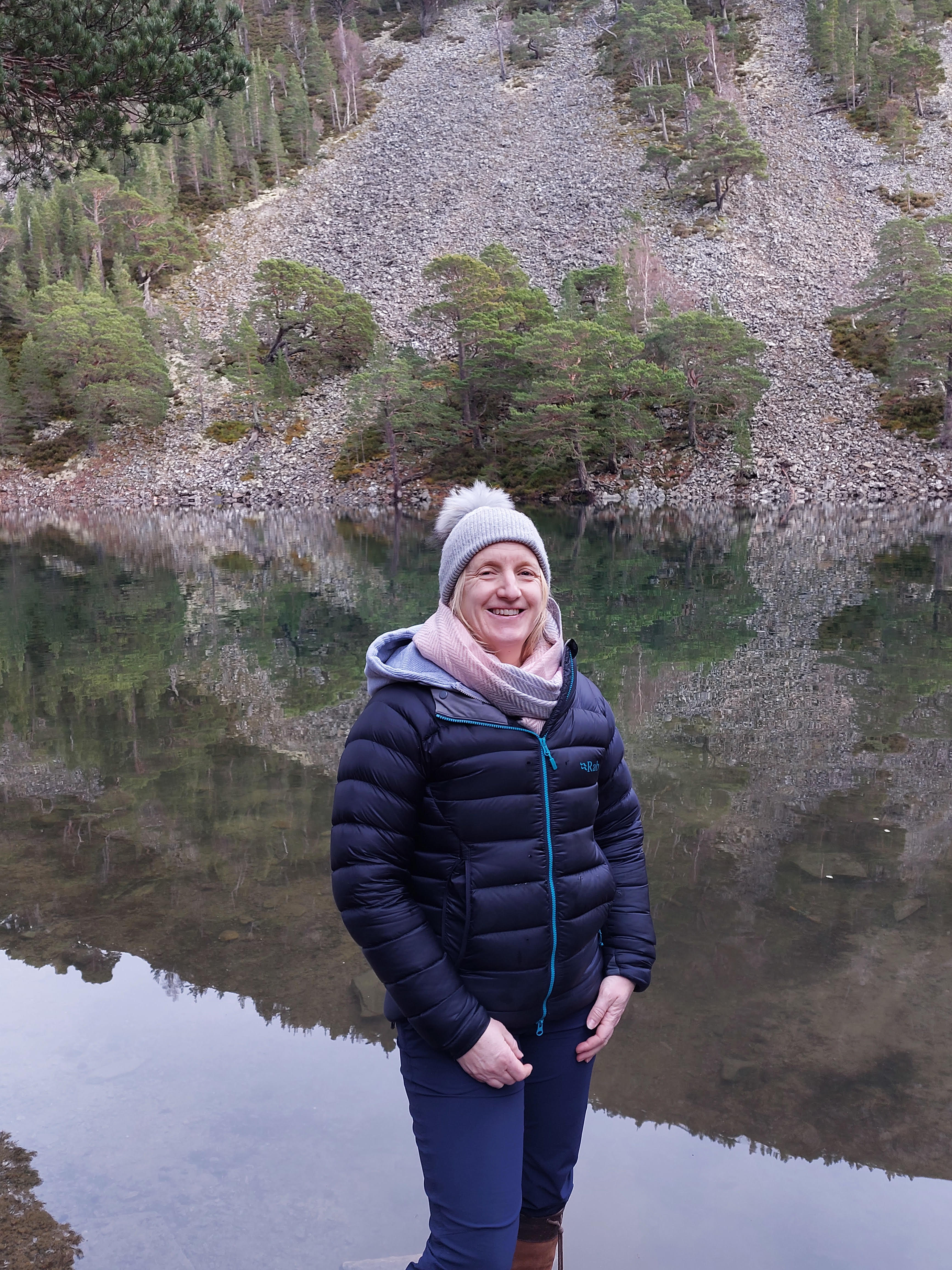 A photograph of Shona in front of a lake. Shona wears a hat and scarf with a puffer jacket and jeans.