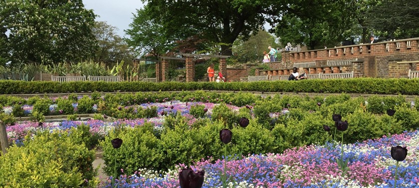 A landscaped garden featuring flowerbeds, trees, and a central walkway. Benches are visible in the background, with people walking and sitting in the area.