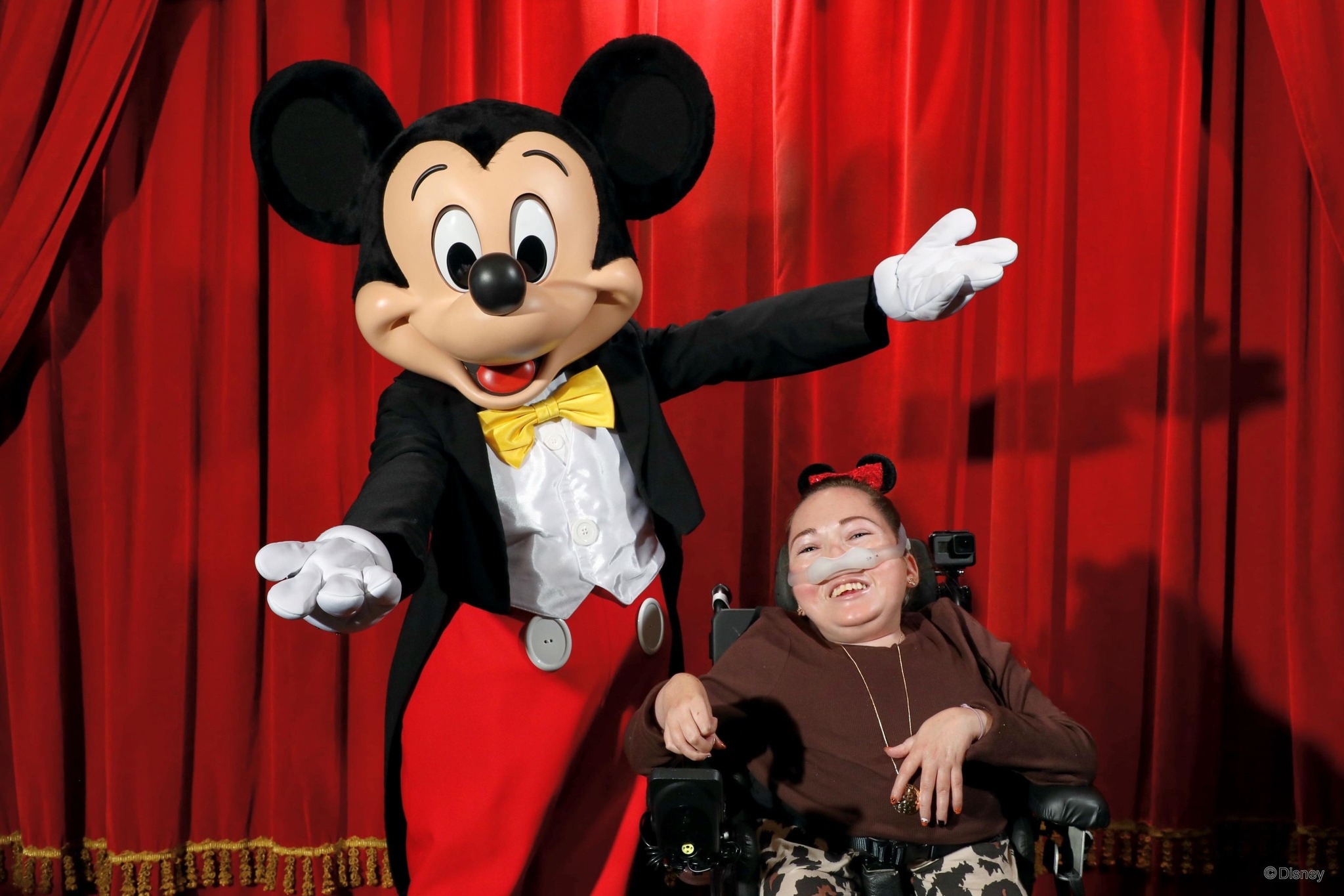 Micky Mouse and Claire pose for a picture in front of a red curtain. Micky has his arms outstretched and Claire is using a powerchair.