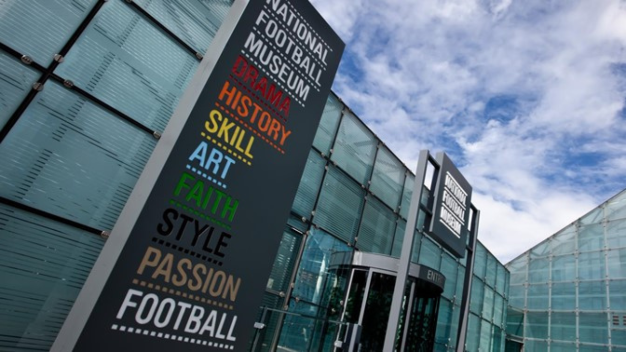 The National Football Museum building, a tall, glass-fronted structure with a triangular shape. The museum name is displayed on the exterior.