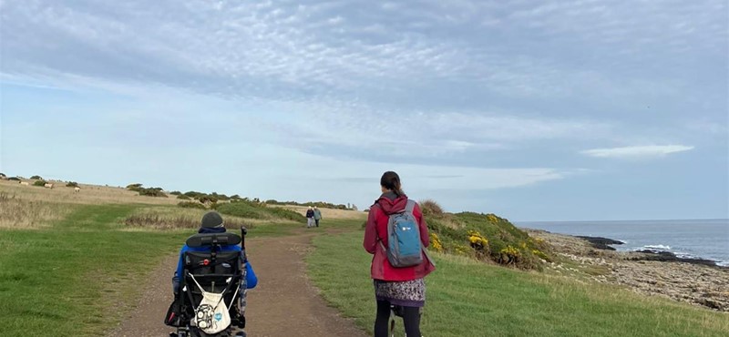 An electric Wheelchair user and a walker wheeling and walking the Craster to Dunsburgh Castle path 