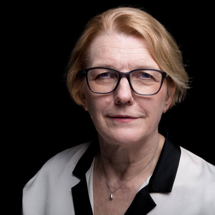 A headshot of Shonaig against a black background. She wears a white top with a black collar and glasses with black frames.