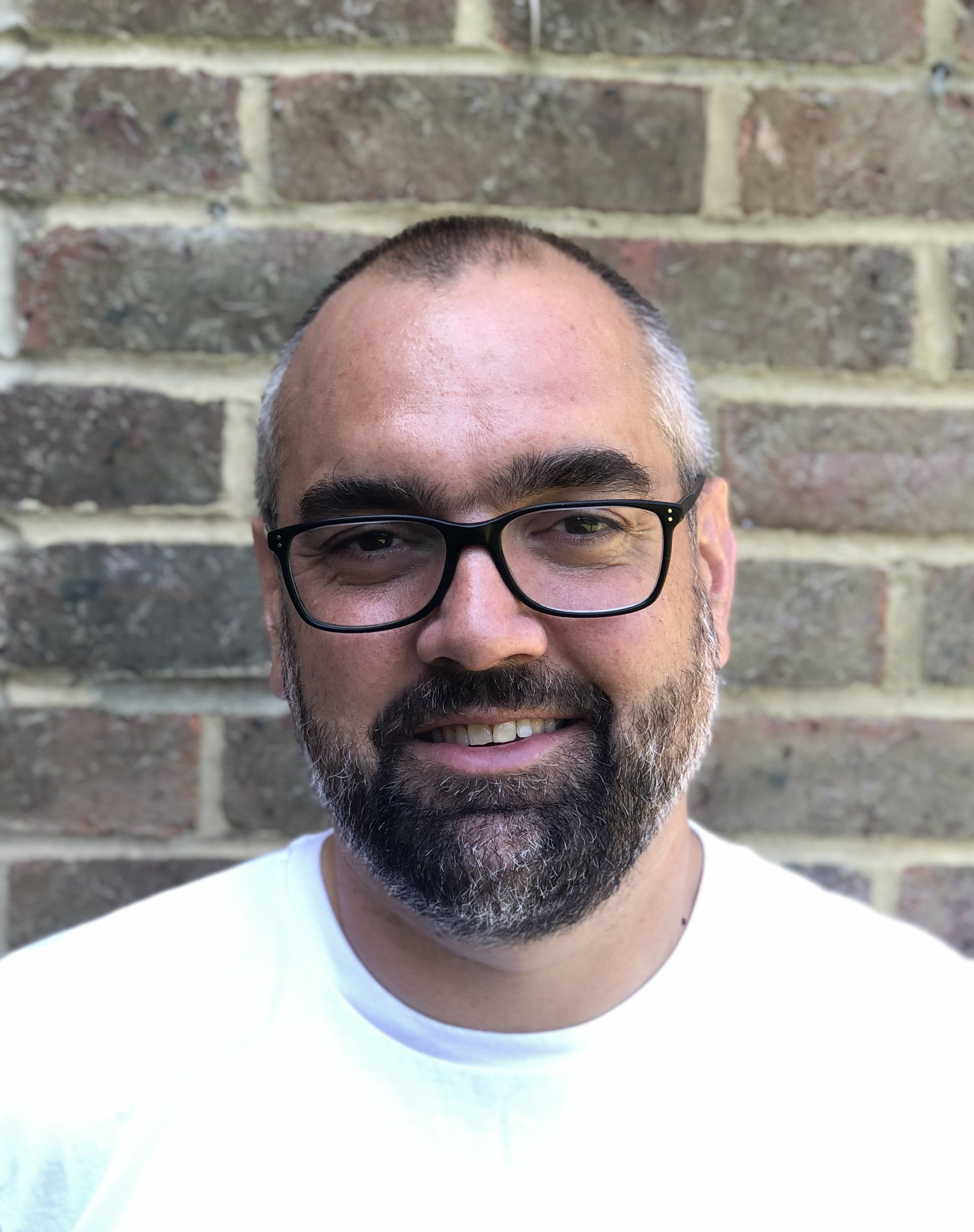 A photograph of Paddy who wears specs, a white t shirt, in front of a brick wall.