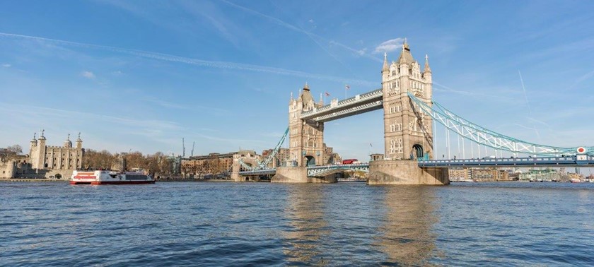 The iconic Tower Bridge with its two tall stone towers connected by a high-level walkway and road deck. The River Thames flows below, with a boat visible on the water.