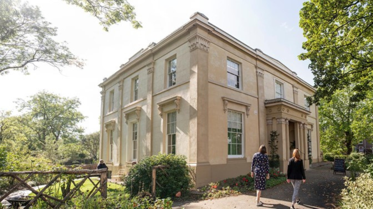 The exterior of Elizabeth Gaskell's House, a two-story Georgian-style building with symmetrical windows and a central entrance. The house is surrounded by a garden with paths and greenery.