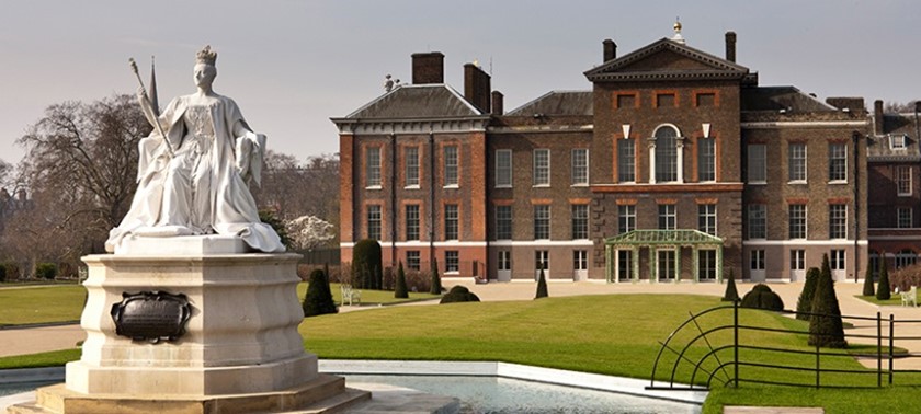 The exterior of Kensington Palace, a red-brick building with large windows and a formal entrance. The surrounding gardens include pathways, open lawns and a large white statue of Queen Victoria.