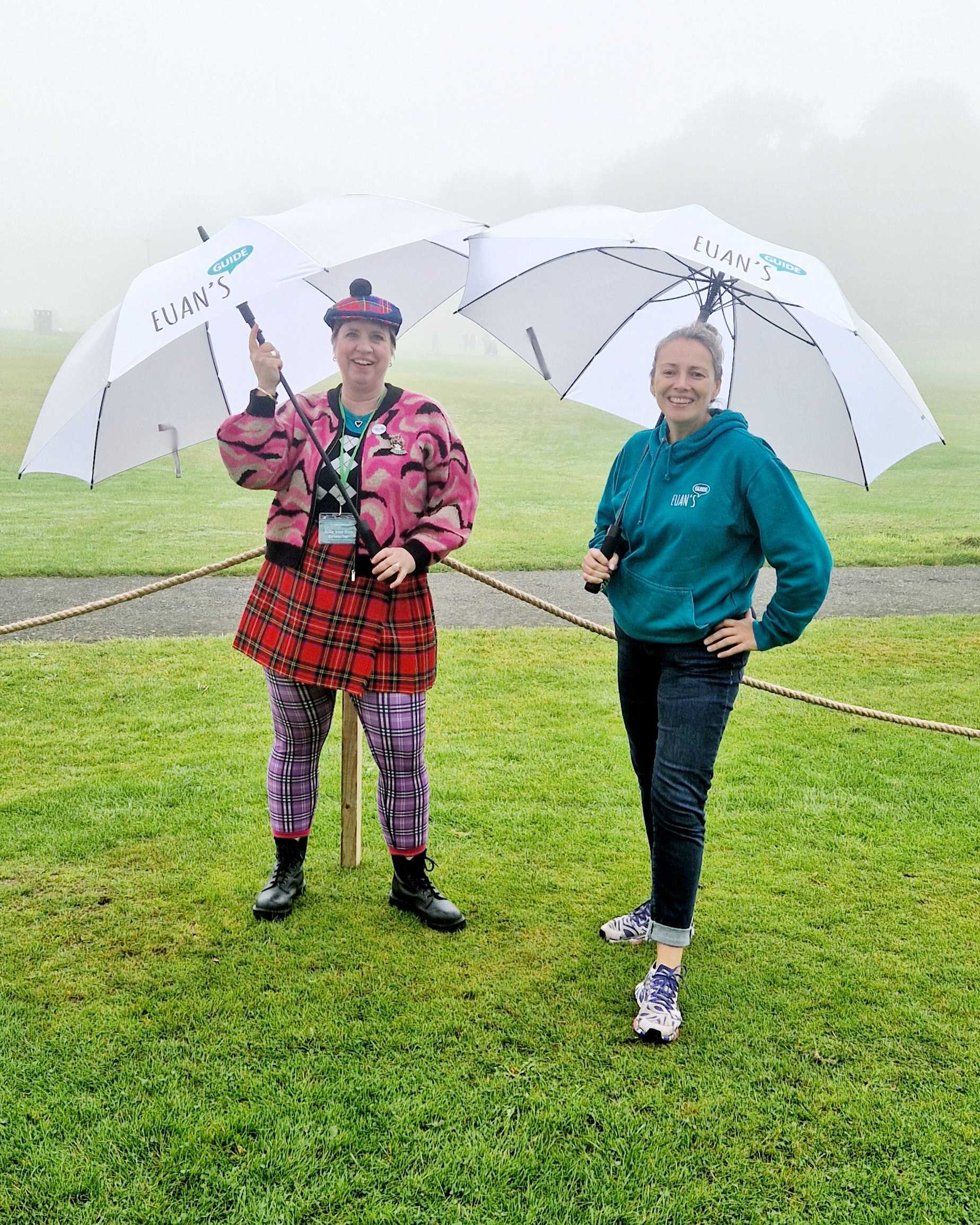 A photo of two women, Julie on the left wearing a tartan skirt and tights with a pink cardigan and Antonia on the right wearing a teal Euan's Guide hoodie, both are holding Euan's Guide golf umbrellas.