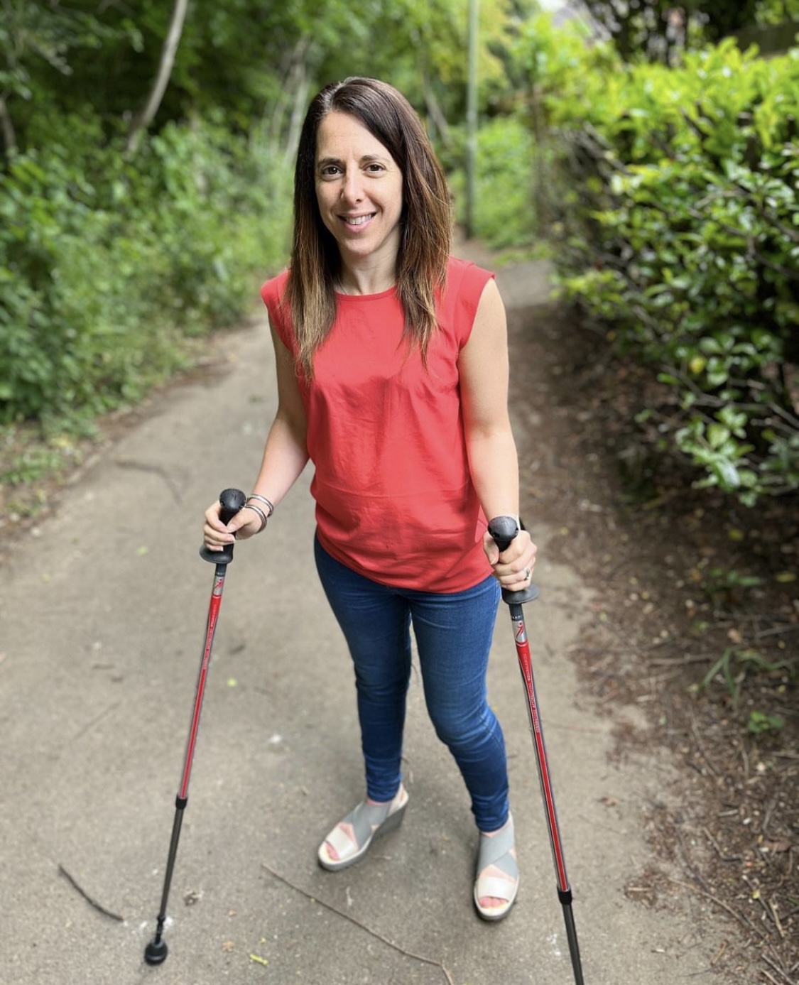 A photo of Georgina wearing a red top and blue jeans and holding walking sticks. Georgina is on a walking /cycle path.