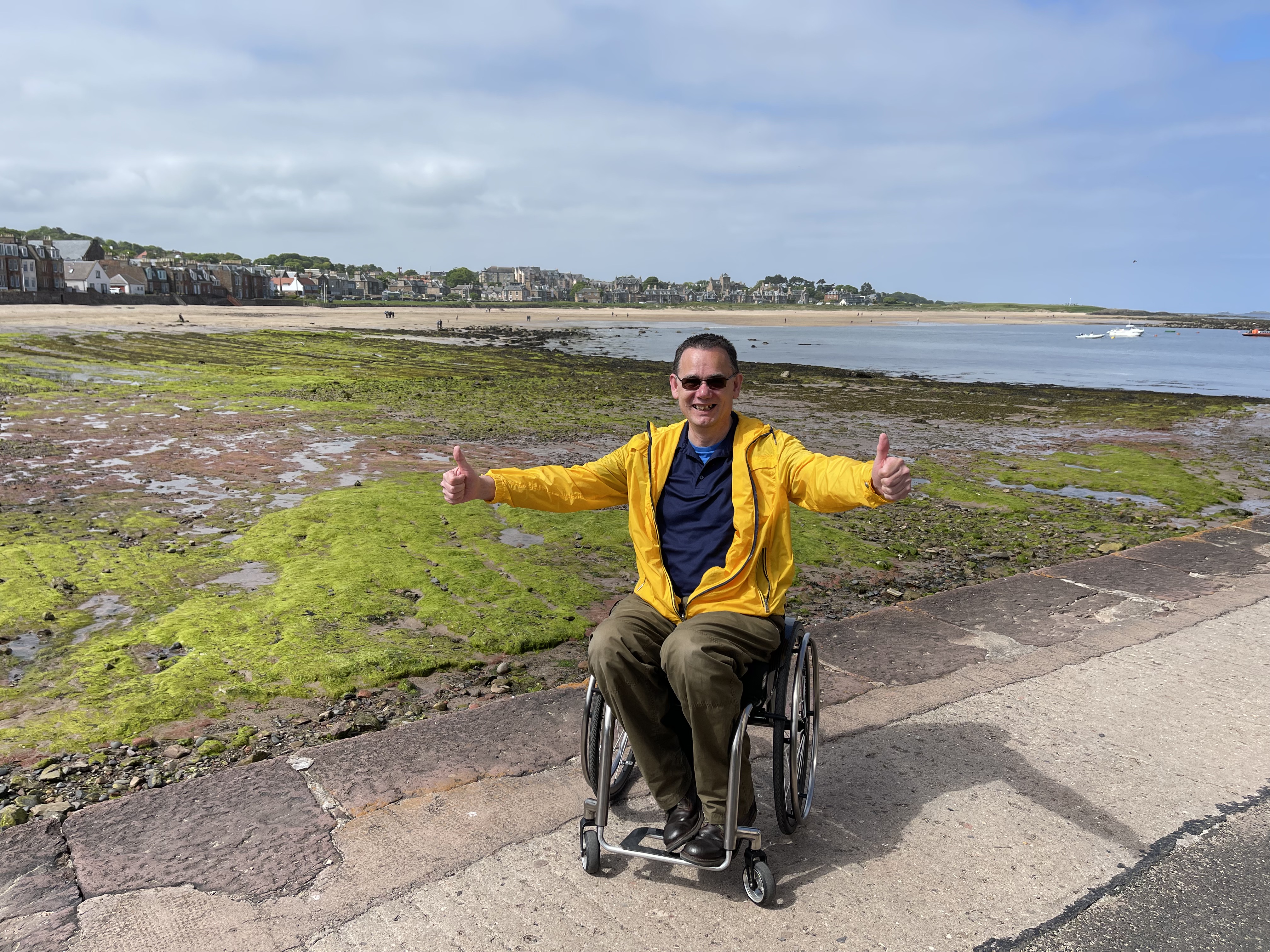 A photograph of Paul at North Berwick harbour, with the tide out in the background. Paul is using a manual wheelchair and  wears a yellow jacket. He's smiling, with his arms outstretched.