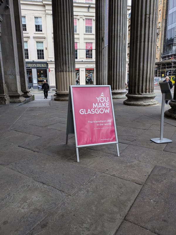 Image of a sandwich board at the entrance, a white stand with a pink poster reading "YOU MAKE GLASGOW"