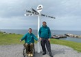 Paul and Ian from Euan's Guide standing alongside the John o' Groats signpost. They are both wearing teal coloured Euan's Guide hoodies.