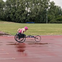 Roz, a white person with long blonde hair is pushing their racing chair. It is a black topend chair (named "The Bimbowagon") with three wheels and stickers on the axel. They are wearing a hot pink helmet. They are racing on an athletics track with red tarmac and white lines. It has just been raining and there are puddles on the track