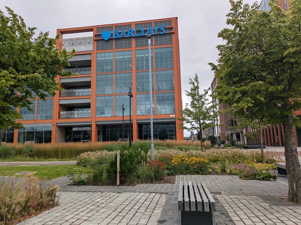 The exterior of the Barclays Campus building, the campus is in the background and a bench and some trees and bushes are in the foreground.