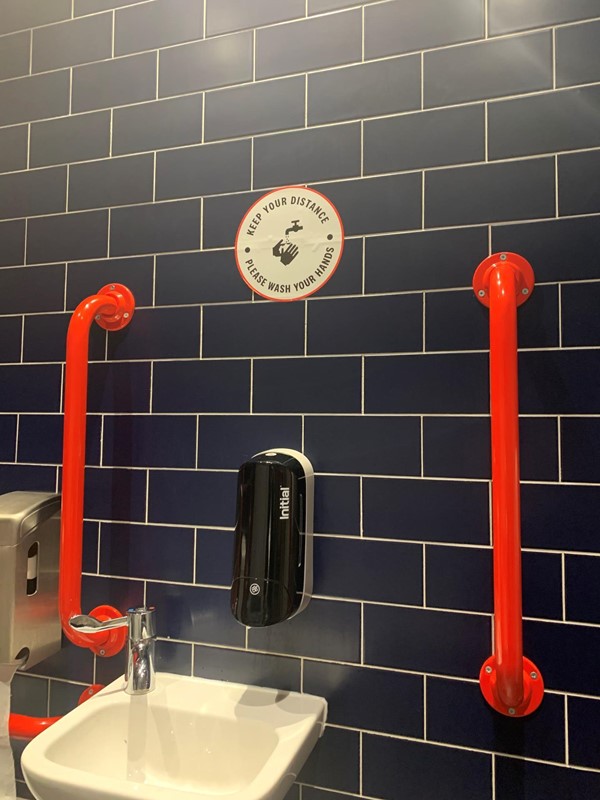 Image of handrails next to sink. There is no mirror but a sticker that says “Keep your Distance, Please Wash Your Hands.”