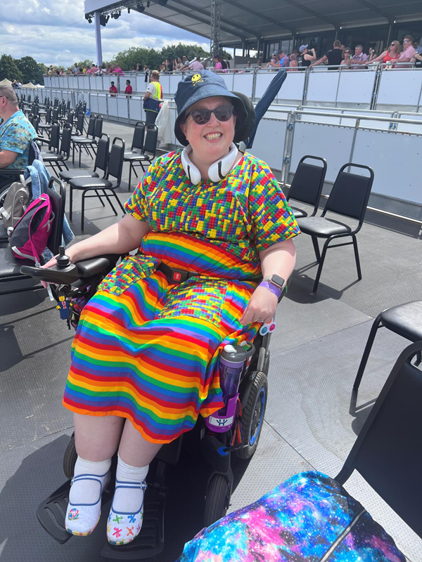 A wheelchair user wearing a rainbow dress, sunglasses and a hat is smiling, they are sitting on the wheelchair viewing platform with VIP areas behind them.