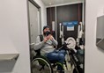 Image of accessible toilet in the mirror. The toilet is reflected as is Joe, a white man wearing a blue hat and grey jumper, he sits on a green wheelchair.