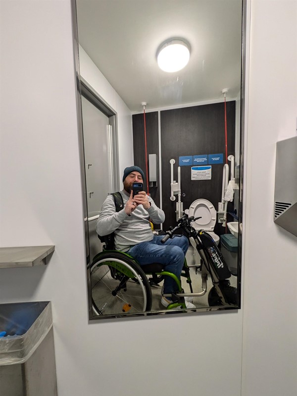 Image of accessible toilet in the mirror. The toilet is reflected as is Joe, a white man wearing a blue hat and grey jumper, he sits on a green wheelchair.