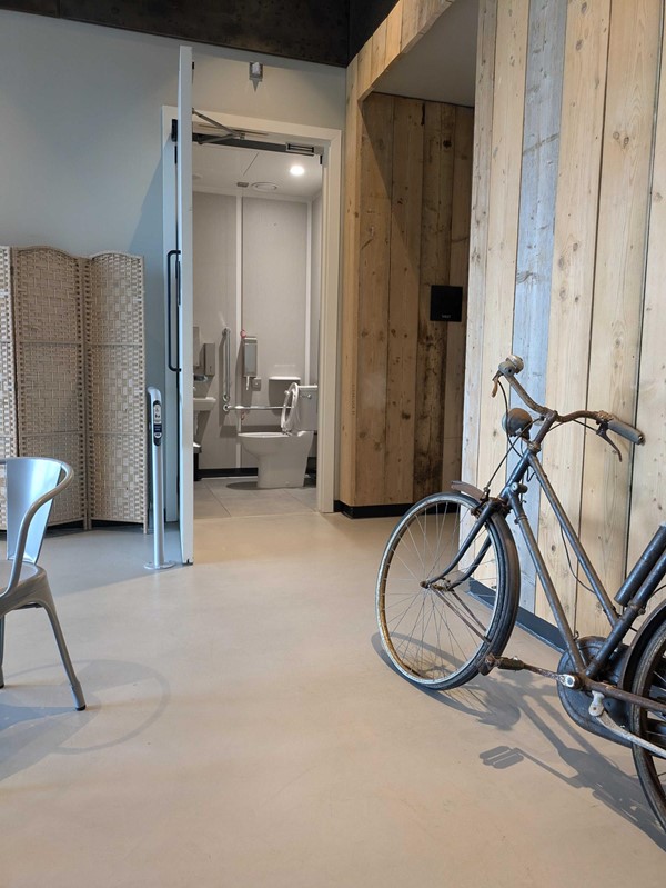 Image of the accessible toilet showing the open door, there's a rusted bike close by and a touchpad outside the door