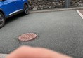 Image of a blue car in a car park with a manhole and a hand