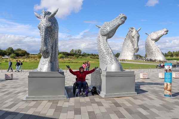 Large flat outdoor area with the Kelpies in the background and two smaller Kelpies on plinths. Lady in wheelchair with a black assistance dog is sitting between the small ones.