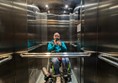 Image of the lift, featuring a mirror and grab rails. Reflected in the mirror i a white man with a shaved head wearing a green hoodie and sitting in a green wheelchair