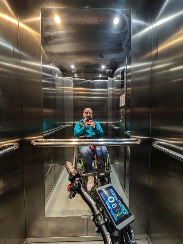 Image of the lift, featuring a mirror and grab rails. Reflected in the mirror i a white man with a shaved head wearing a green hoodie and sitting in a green wheelchair