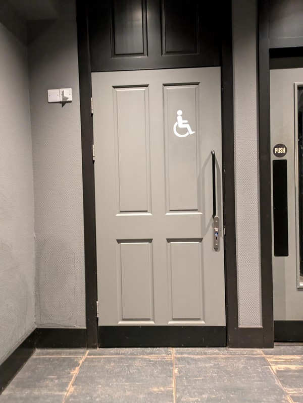 Entrance to the accessible toilet. A grey door with a white wheelchair user symbol on the top right.