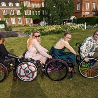 a group of four manual wheelchair users in a line, holding on to each others backrests and sitting sideways to the camera. They all have decorated their wheels in different ways. Els (far right) has rainbow spokes. Katy (right) has purple wheels with purple and black spokes. Roz (left) has pink cow print wheel covers. Holly (Far left) has red wheels with multicoloured spokes. They are all smiling at the camera