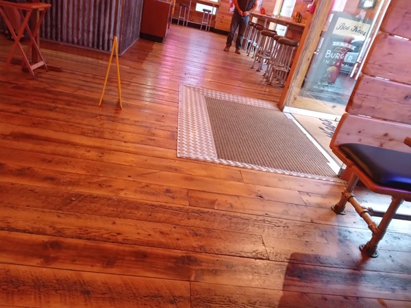 a photograph of the entrance from the inside. just inside the door there is a fixed doormat and a small metal slope. the rest of the floor is smooth floorboards. table legs are visible around the outside of the frame.