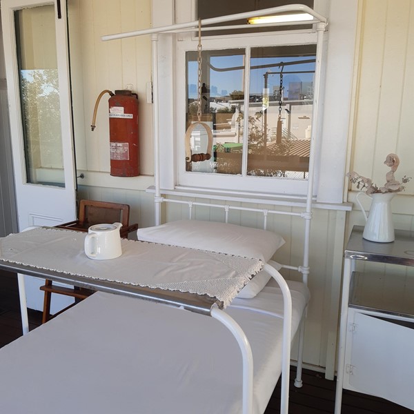 There is an old hospital bed on a verandah that has two white pillows and a white sheet on it. There is a small table that is over the bed. The table has a white doily on it. There is a white teapot on the table. End Description.