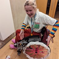 Roz has just discovered what happens if you take a wheelchair in the snow. They are sitting in a grey hospital chair with pink spoke covers on the wheels. the entire push rim is coated in snow. they are gesturing at the wheel and have an expression on their face which reads as "what am i meant to do with this?".