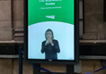 Image of a BSL Departure Board - a vertical black screen showing a white woman with blonde hair signing