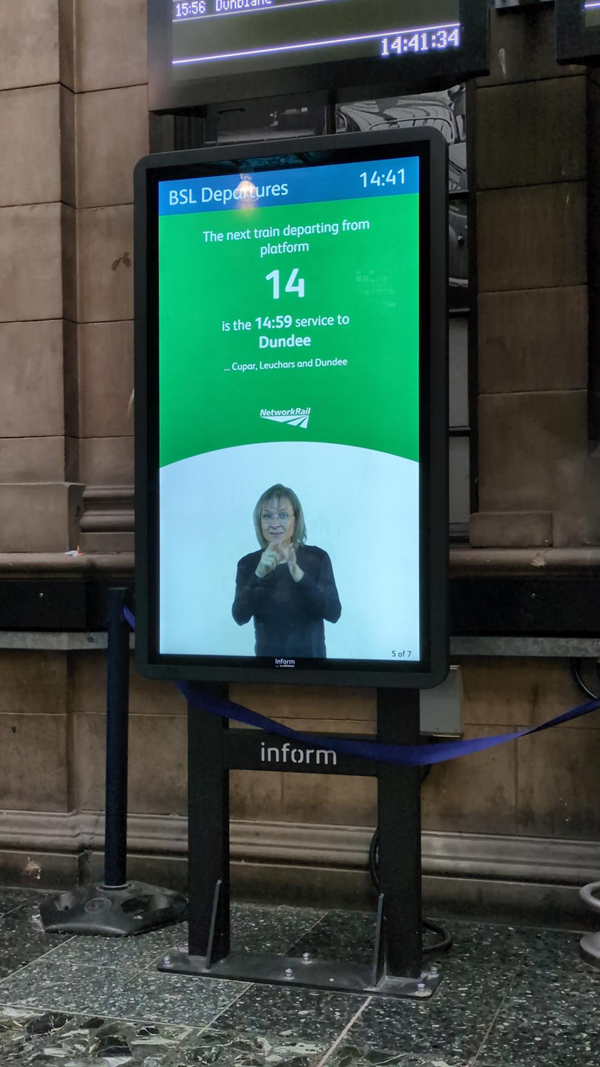 Image of a BSL Departure Board - a vertical black screen showing a white woman with blonde hair signing