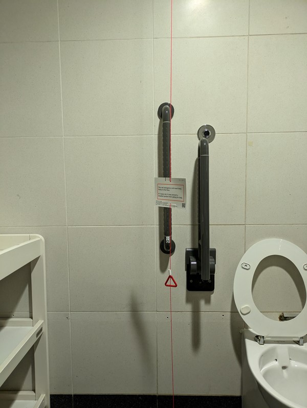 Image of a toilet with grab rails, the red cord has a red cord card attached and now touches the ground