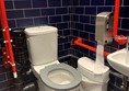 Image of accessible toilet showing handrails, toilet, red cord card hanging behind the bin and sink.