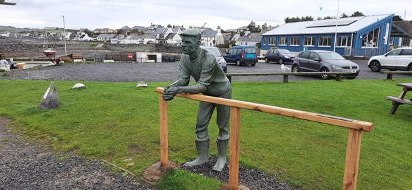Statue of a man in a flat cap leaning against a wooden fence