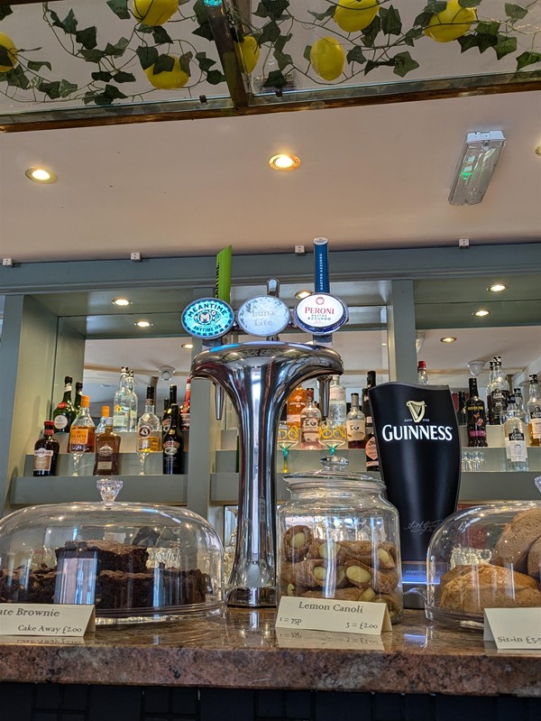 Image of the beer taps, the labels of which read Meantime IPA, Luna Lite, Peroni and Guinness. There are cakes on the bar top, brownies, cannolis and scones