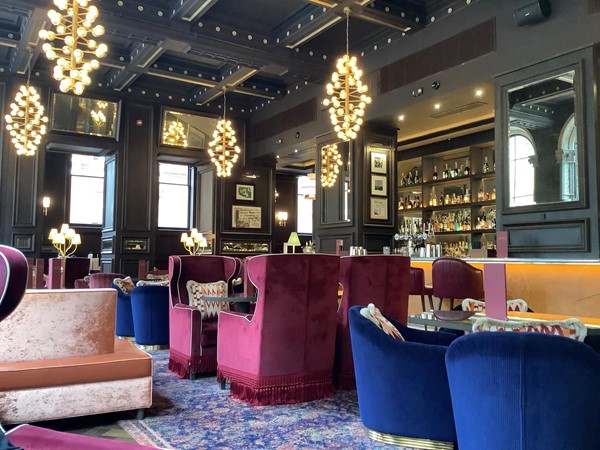 Interior of hotel featuring high back red velvet sofas and low navy blue velvet tub chairs with a blue carpet and dangling lighting