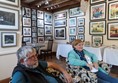 Elderly couple sitting in armchairs, with framed pictures on the walls behind them.