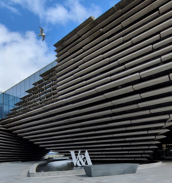 Exterior of the V&A, a white stylised V&A sign, the building is large and grey with a corregated appearance.