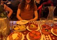 Photograph shows a young wheelchair user at a table with lots of tasty Tapas in front of her. Behind her is another table with a number of wheelchair users sat comfortably