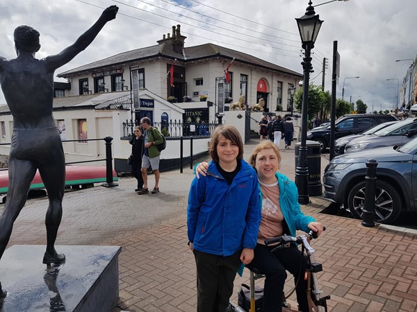 LondonMarmot on small scooter on the promenade next to the sea in Cobh. The Titanic Experience is in the old White Star Line building in the background. The statue on the left is of Irish athletics champion Sonia O'Sullivan