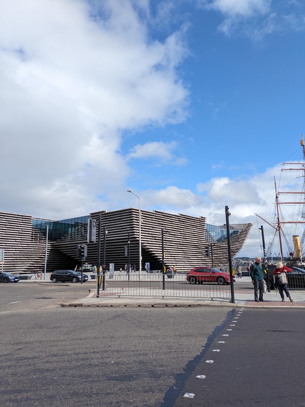 Image of the road crossing at the V&A. Two people are waiting to cross, the V&A is in the background and the mast of the RRS Discover is in the background.