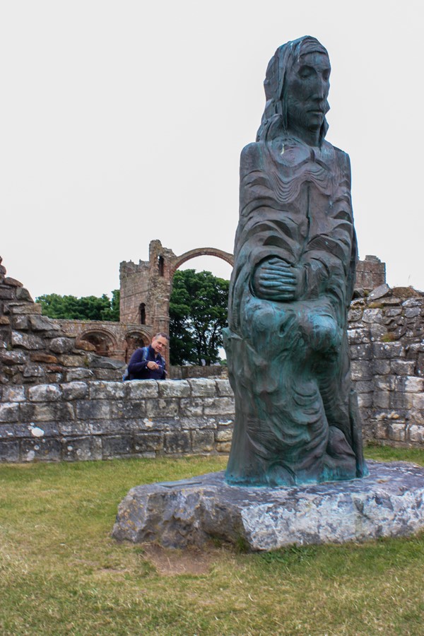 Statue of Cuthbert standing on the grass and ruins in the background.